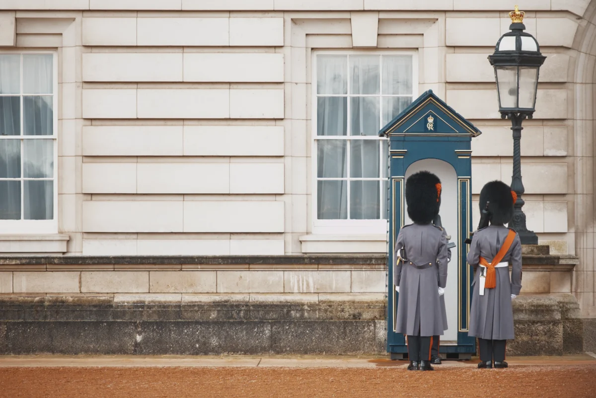 Photographie d'art minimaliste de deux gardes de la reine (Coldstream Guards) de dos devant une guérite bleue au palais de Buckingham à Londres, grand espace vide en pierre à gauche.