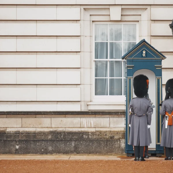 Photographie d'art minimaliste de deux gardes de la reine (Coldstream Guards) de dos devant une guérite bleue au palais de Buckingham à Londres, grand espace vide en pierre à gauche.