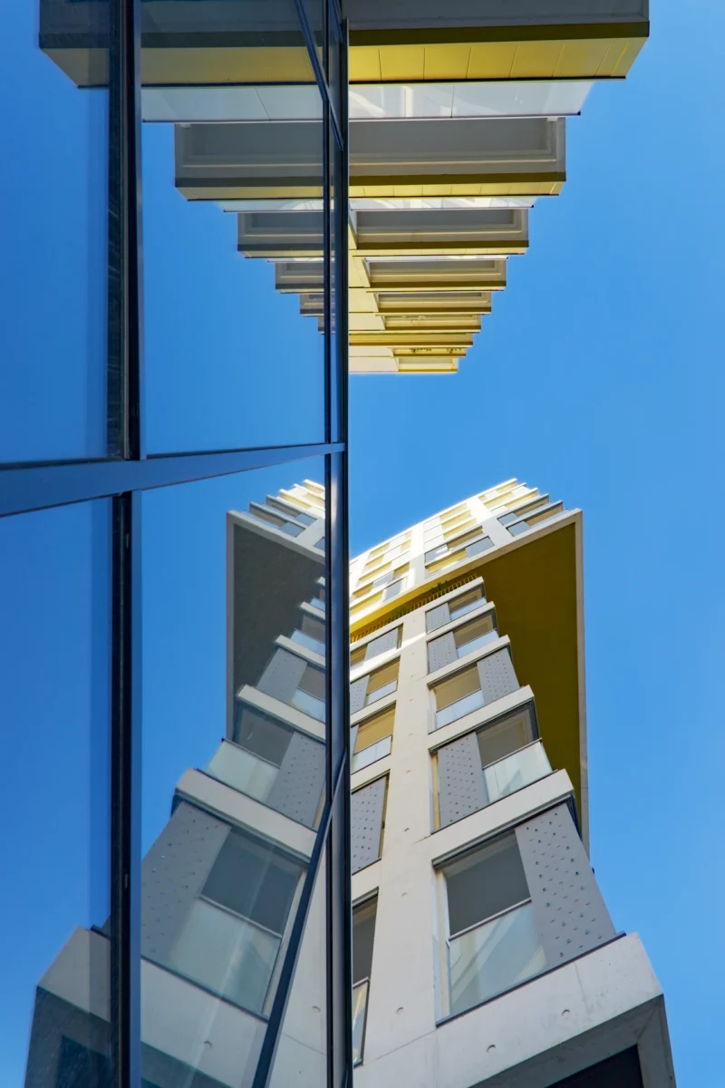 Photographie d'art en contre-plongée de la résidence Palomaya, montrant la façade blanche et or se reflétant symétriquement dans une paroi de verre noire face à un ciel bleu.