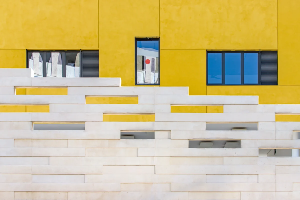 Joan Miro est une photographie d'art minimaliste d'une façade d'école jaune avec un mur blanc en forme de tétris, des fenêtres reflétant le ciel bleu et une pastille rouge au centre.