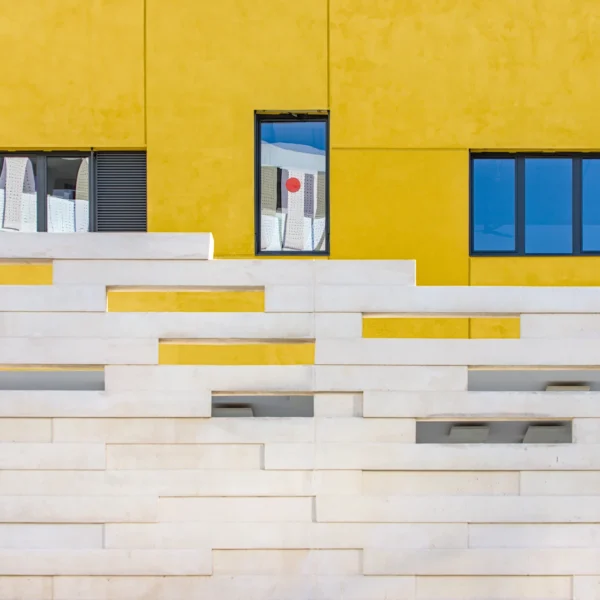 Joan Miro est une photographie d'art minimaliste d'une façade d'école jaune avec un mur blanc en forme de tétris, des fenêtres reflétant le ciel bleu et une pastille rouge au centre.
