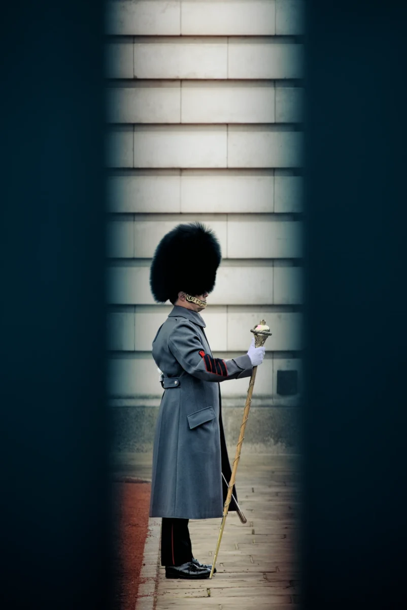 Maestro Buckingham est une photographie d'art d'un garde royal de Buckingham de profil en capote grise et bonnet noir, tenant son bâton de commandement, cadré entre deux barreaux sombres en hiver.