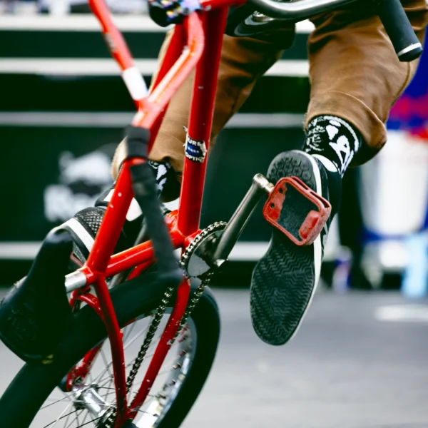 Gros plan artistique d'une fusion mécanique et corporelle : pied d'un rider en tension sur la pédale d'un BMX au cadre rouge, triade de couleurs au FISE Montpellier.