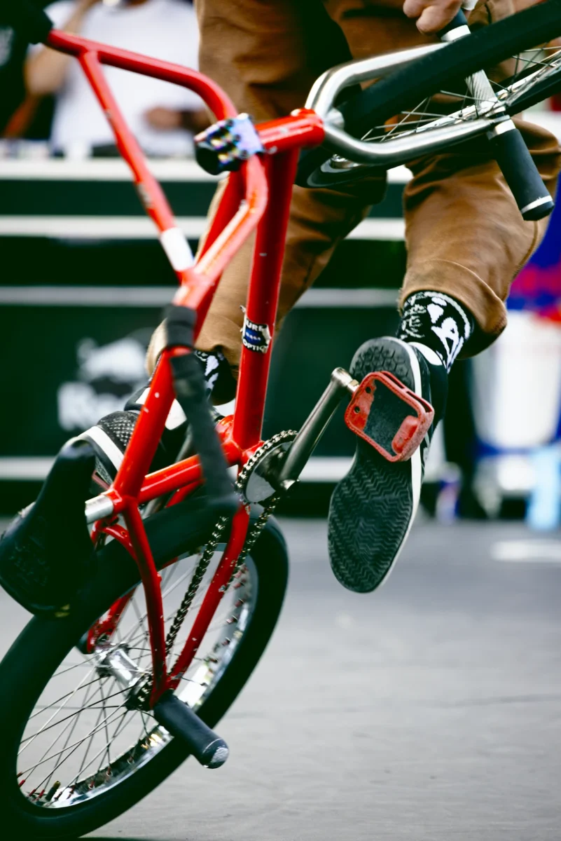 Gros plan artistique d'une fusion mécanique et corporelle : pied d'un rider en tension sur la pédale d'un BMX au cadre rouge, triade de couleurs au FISE Montpellier.